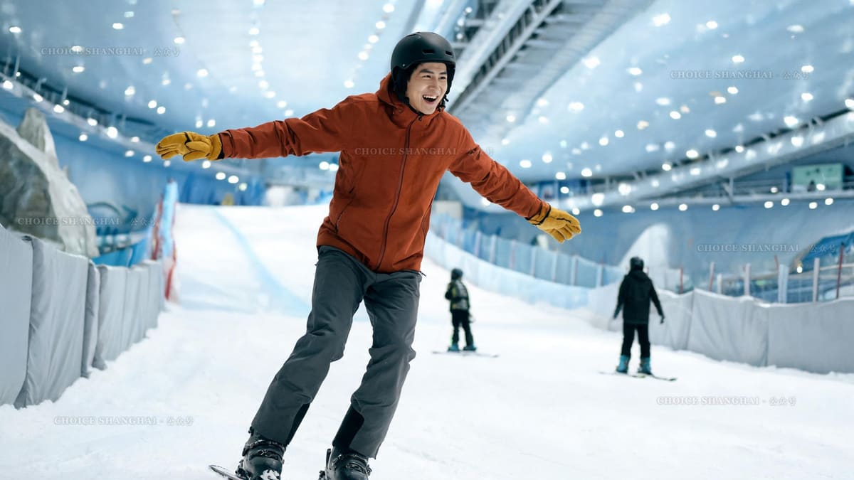 First timer laughing mid wobble on a beginner slope inside an indoor snow hall, arms out wide in a rust orange jacket and mustard gloves