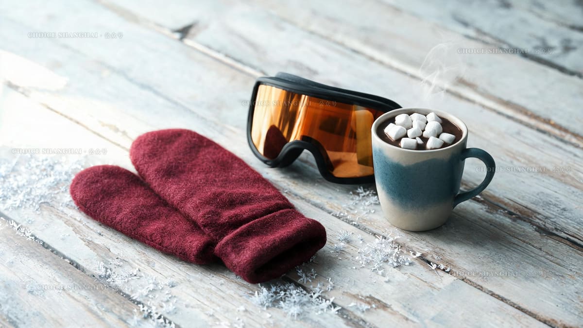 Burgundy wool mittens, amber ski goggles and a mug of hot cocoa with marshmallows on pale weathered wood, frost scattered around