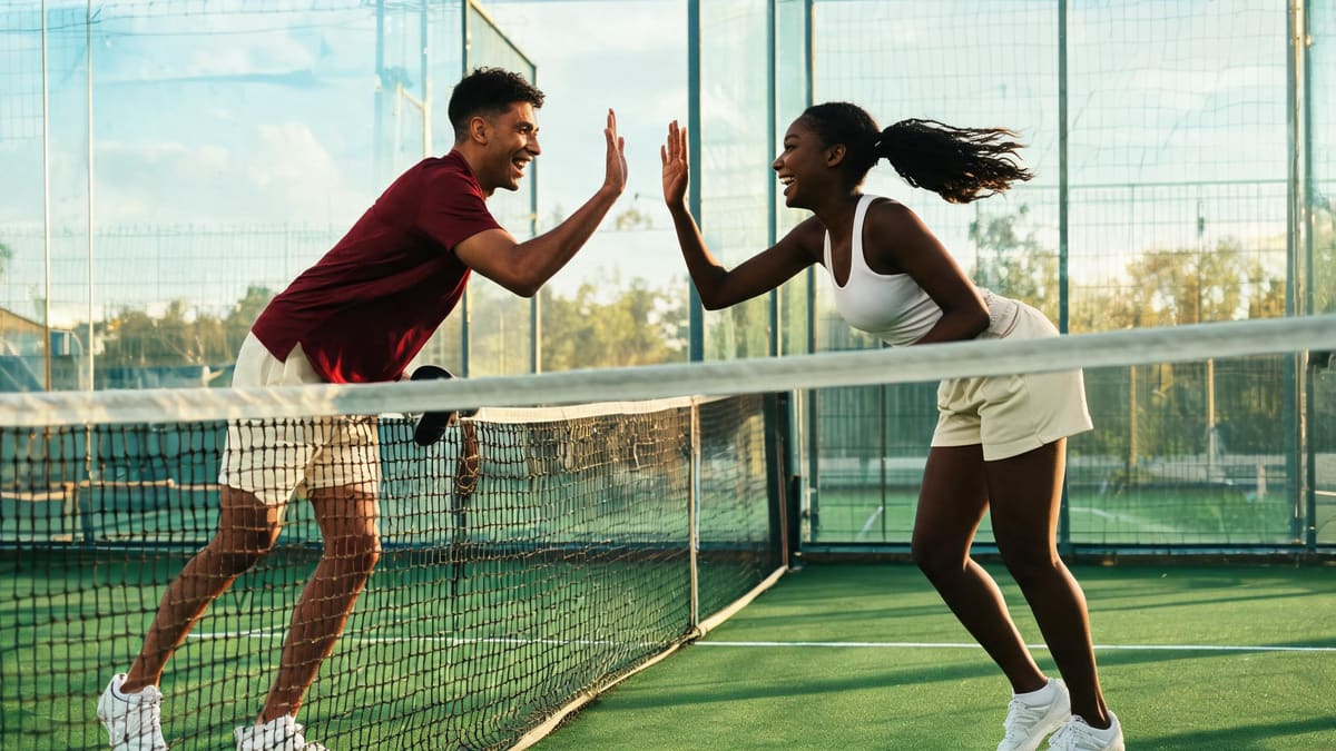 Two friends in summer sportswear high-fiving over the net on a glass-walled padel court in Shanghai