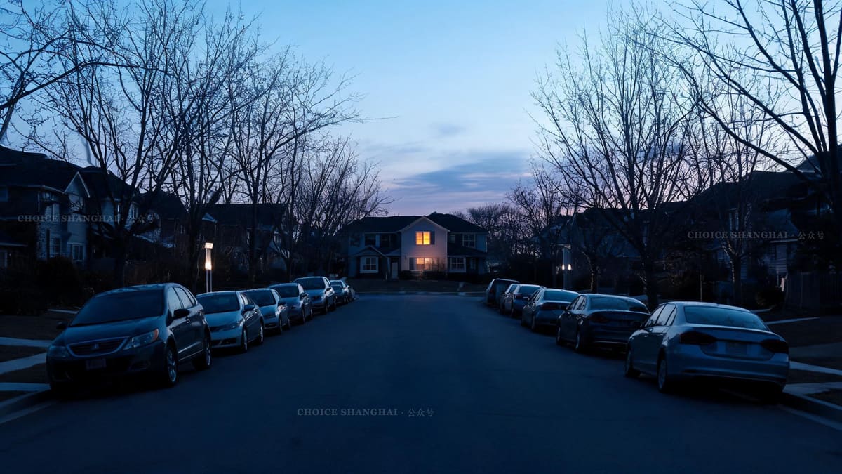 A very quiet suburban street at dusk in a Western town, empty pavements and still trees, a single lit window in the distance