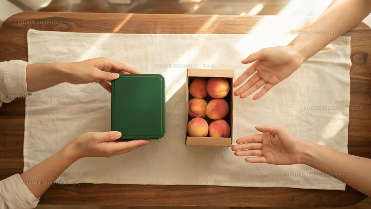 Two pairs of hands exchange a green tea tin and a kraft box of fresh peaches across a linen table runner in soft light