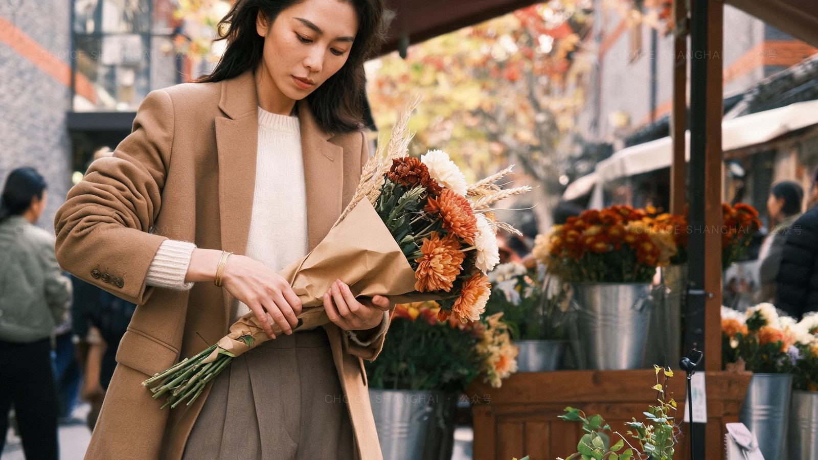 A woman in a camel coat wrapping a bouquet of autumn chrysanthemums in brown paper at a Shanghai flower-market stall