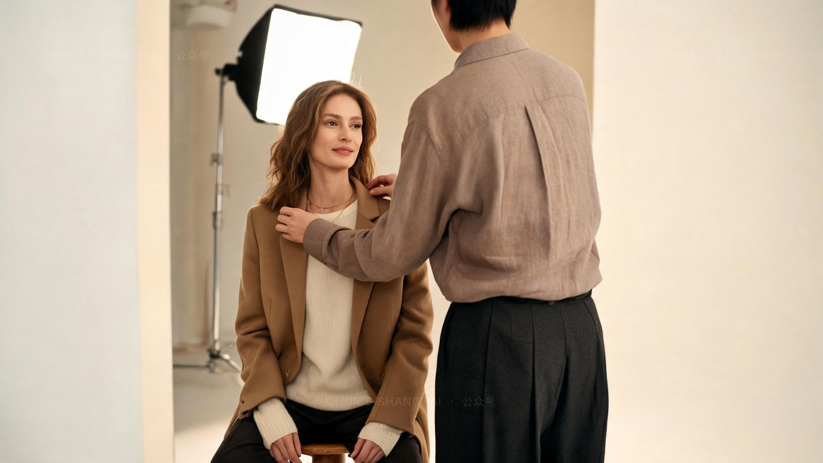 A woman in a camel blazer seated on a stool under a studio softbox in a Shanghai photo studio while a stylist adjusts her collar