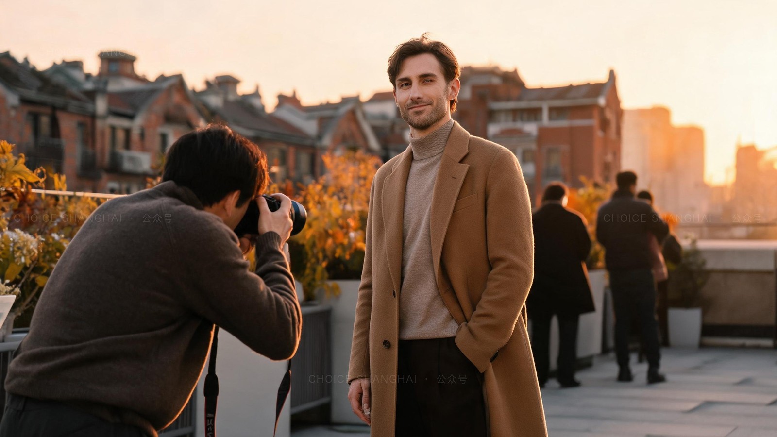 A man in a camel overcoat being photographed on a Shanghai rooftop at golden hour, the photographer kneeling in the foreground, old brick rooftops behind