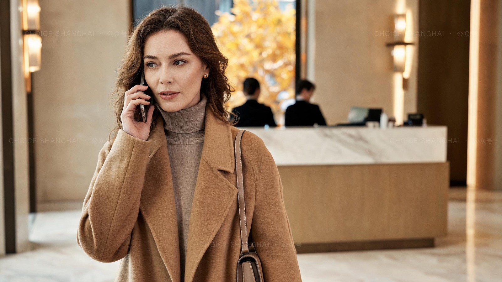 A woman in a camel coat in a residential building lobby talking on her phone, a property-management reception desk softly out of focus behind her, warm light