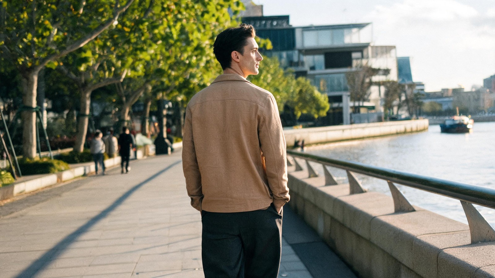 A man walking alone along the West Bund riverside promenade in Xuhui on a clear autumn afternoon, water and art museums beside him