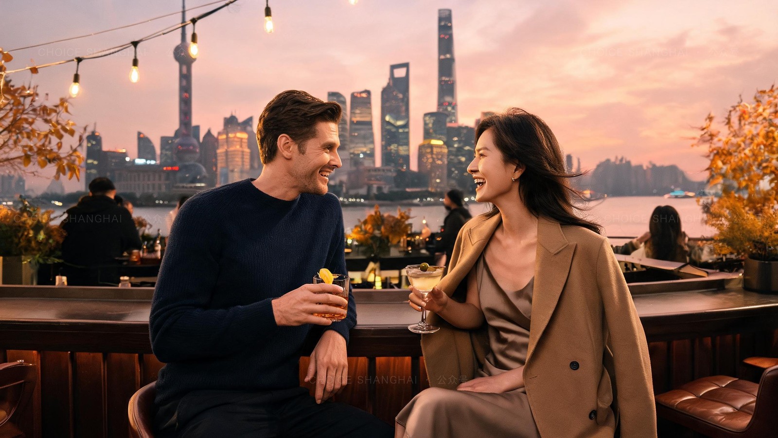 A couple laughing over cocktails at a Bund-facing rooftop bar at sunset, the lit Pudong skyline across the river behind them
