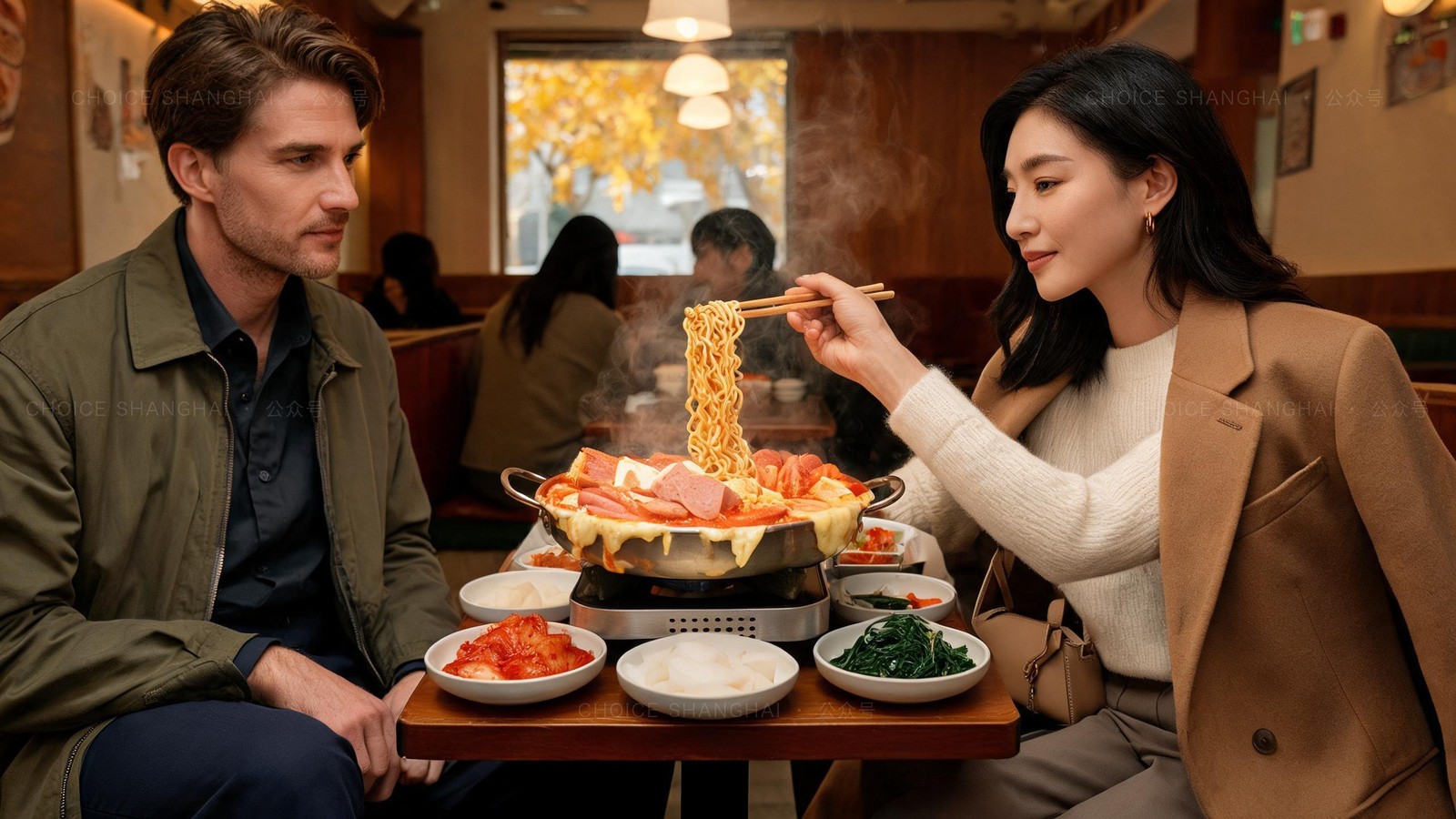 Bubbling Korean army stew in a metal pot on a tabletop burner, with sausage, tofu, cheese and instant noodles being lifted with chopsticks, banchan bowls around it