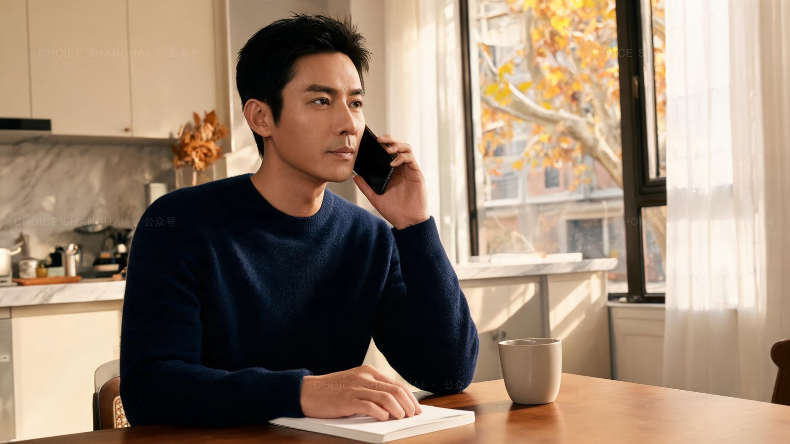 A man in a navy sweater making a composed phone call at his kitchen table in a Shanghai apartment, notebook open, autumn window light