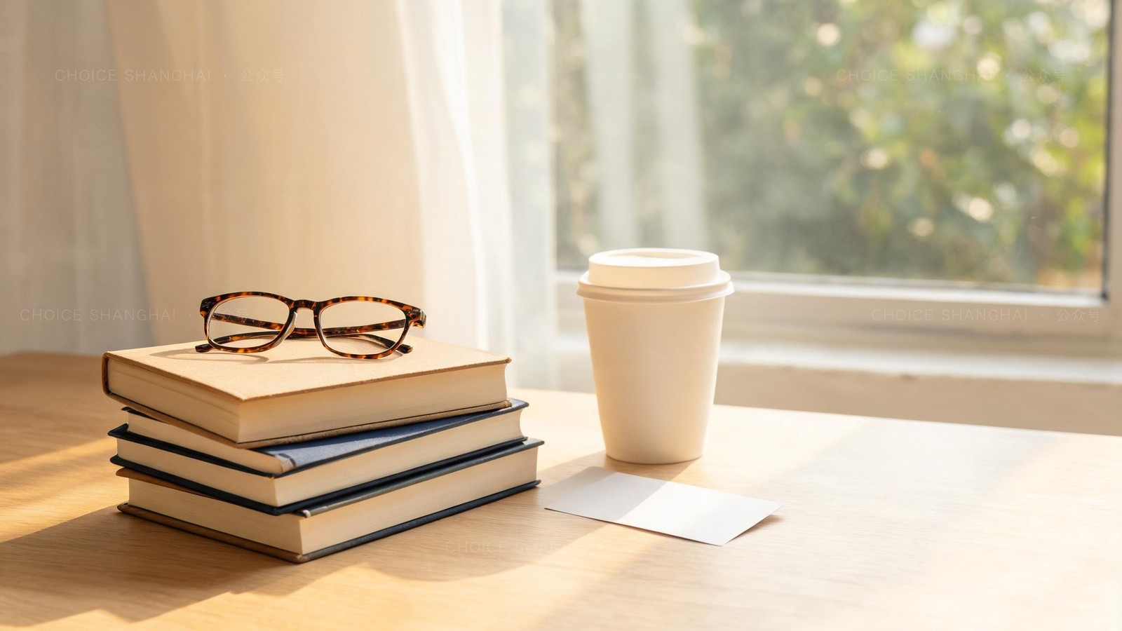 A stack of hardcover books with tortoiseshell reading glasses on top, a takeaway coffee and a blank library card on a sunlit wooden table