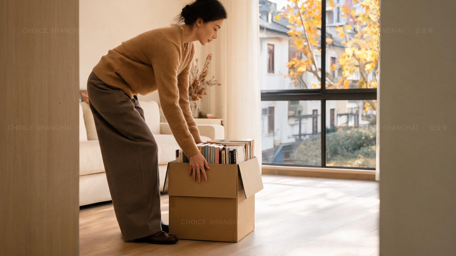 A woman in a camel sweater setting down a box of books in a bright new Shanghai apartment with golden autumn leaves outside the window