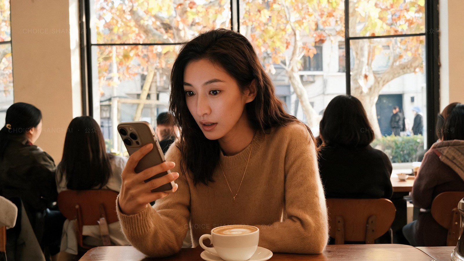 A woman in a camel knit at a sunlit Shanghai cafe looking at her phone with a wry, surprised expression, autumn leaves outside the window