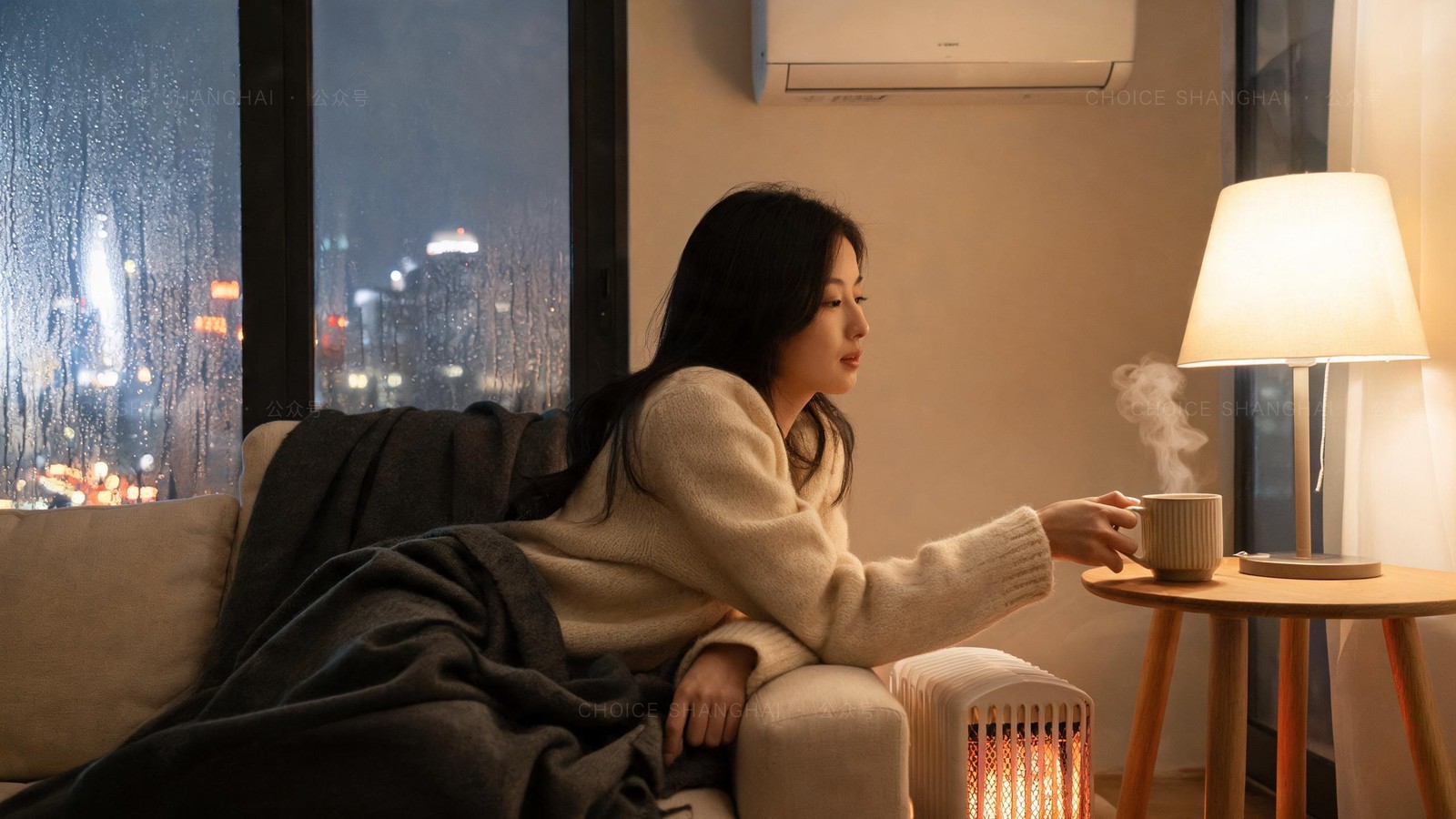 Woman in a cream cashmere knit reaching for a steaming mug beside an oil heater and reverse-cycle air conditioner in a Shanghai flat on a rainy winter night